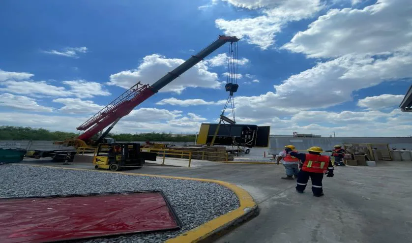 Grúa roja izando un equipo en el patio de una planta, con personal en chalecos de alta visibilidad y área delimitada