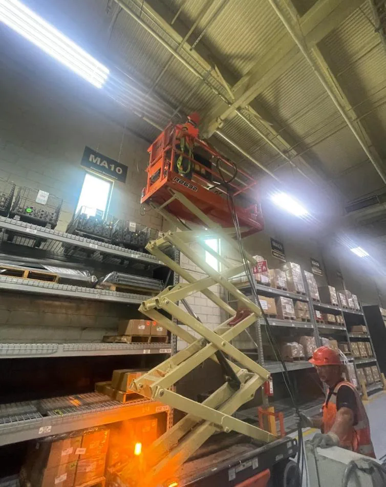 Plataforma de tijera extendida a gran altura en un pasillo de almacén entre racks, con técnico con casco naranja